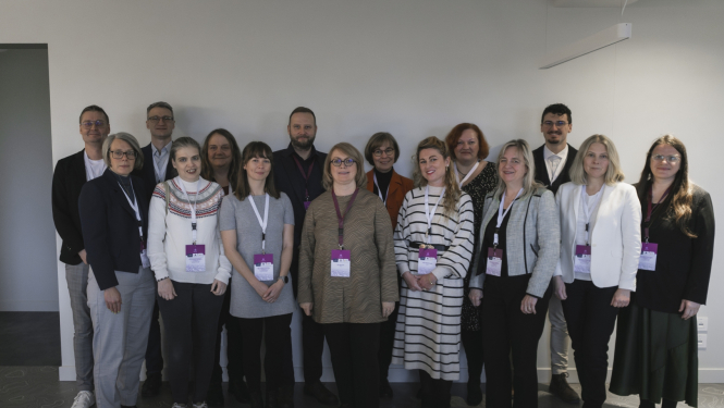 Konferences dalībnieku foto gaišā telpā, kurā redzami dažādu iestāžu pārstāvji. Visi dalībnieki stāv rindās pie sienas, valkā pasākuma identifikācijas kartes un oficiālu vai biznesa ikdienas apģērbu, radot organizētu un formālu noskaņu.