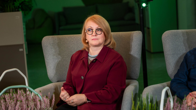 Valsts zemes dienesta ģenerāldirektore sēž krēslā , tērpusies tumši sarkanā žaketē. Priekšplānā redzami dekoratīvi augi, bet fonā — gaišs interjers ar dīvāniem un apgaismojumu.