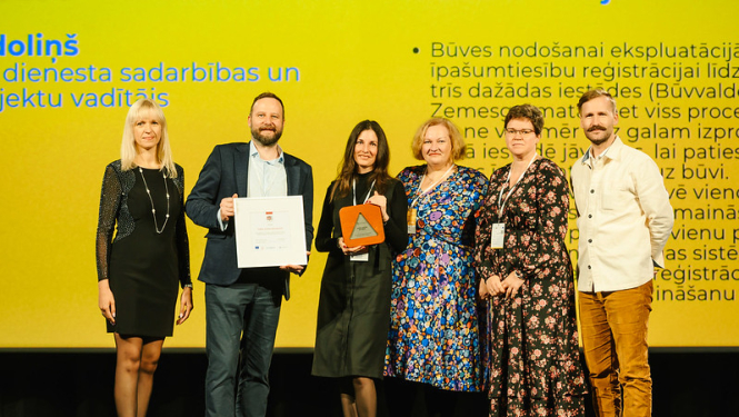 Sešu cilvēku grupa stāv skatuves priekšā pasākumā, rokās turot apbalvojumus un sertifikātu. Fonā redzams liels ekrāns ar dzeltenu prezentācijas slaidu un tekstu par sadarbību un būvju reģistrācijas procesu.
