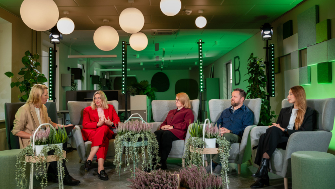 Paneļdiskusijas dalībnieku foto telpā ar zaļiem gaismas akcentiem. Dalībnieki sēž mīkstos krēslos puslokā, starp viņiem izvietoti dekoratīvi augi. Fonā redzams moderna interjera dizains ar griestu lampām, sienu paneļiem un telpaugiem.