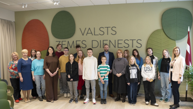 Grupas foto Valsts zemes dienesta telpā pie sienas ar VZD nosaukumu un stilizētiem koku lapu dekoriem. Priekšplānā redzami Valsts zemes dienesta darbinieki un jaunieši, kuri piedalās "Ēnu diena" pasākumā. Fonā redzams arī Latvijas karogs un moderna, gaiša telpa.