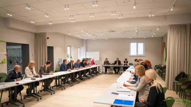 Gaiša sanāksmju telpa, kur pie divām galdų rindām profesionālā darba apspriedē piedalās pārstāvji no Valsts zemes dienesta un Saeimas Valsts pārvaldes un pašvaldības komisijas. Priekšgalā sēž prezentētāji, telpā moderns apgaismojums un neitrāls interjers.
