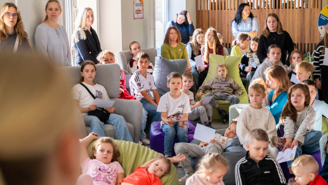 Pasākuma dalībnieki pulcējušies gaišā telpā uz kopīgu nodarbību. Daži sēž uz pufiem vai dīvāniem, citi stāv, rokās turot papīrus. Telpā redzams silts apgaismojums un koka interjera elementi.