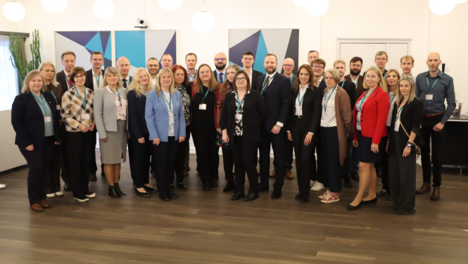 Pasākuma dalībnieku kopējā fotogrāfija iekštelpās konferenču vidē. Dalībnieki ir dažādos profesionālos tērpos un ar vārdu kartītēm, fonā redzami ģeometriski sienu dekori un telpas interjers.