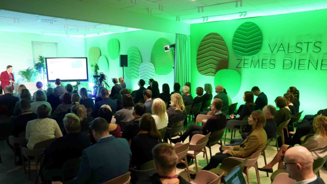 Valsts zemes dienesta Kadastra zāles foto, kurā notiek prezentācija par aktualitātēm. Dalībnieki sēž rindās, klausoties runātāju pie ekrāna. Telpu izgaismo spilgta zaļā gaisma un sienas rotā stilizēti koku lapu paneļi ar VZD nosaukumu, radot dinamisku un mūsdienīgu vidi.