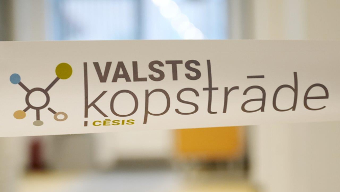 Tuvplāna attēls ar balti laminētu lentu, uz kuras redzams “Valsts kopstrāde Cēsis” vizuālais apzīmējums un stilizēta sadarbības tīkla ikona. Fons ir gaišs biroja gaiteņa interjers ar izpludinātām formām, kas izceļ priekšplānā redzamo zīmolu.