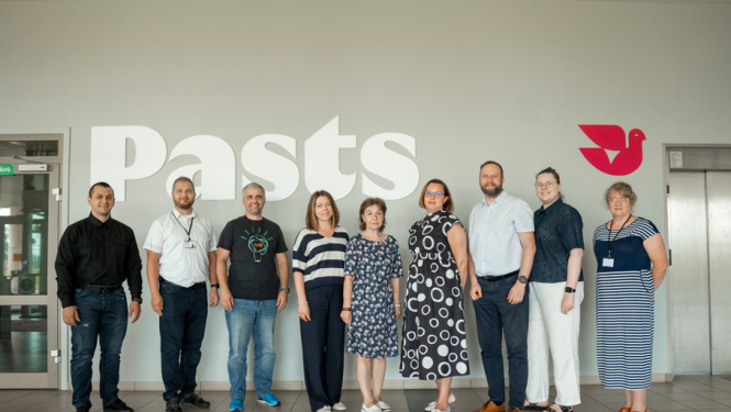 Tikšanās dalībnieki pie Latvijas Pasta uzraksta un zīmes, stāvot kopā ēkas vestibilā.