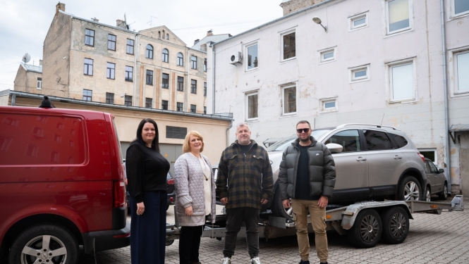 Četri cilvēki stāv pagalmā pie sarkanas furgona un piekabes ar sudraba apvidus automašīnu; fonā redzamas vecākas daudzdzīvokļu ēkas.