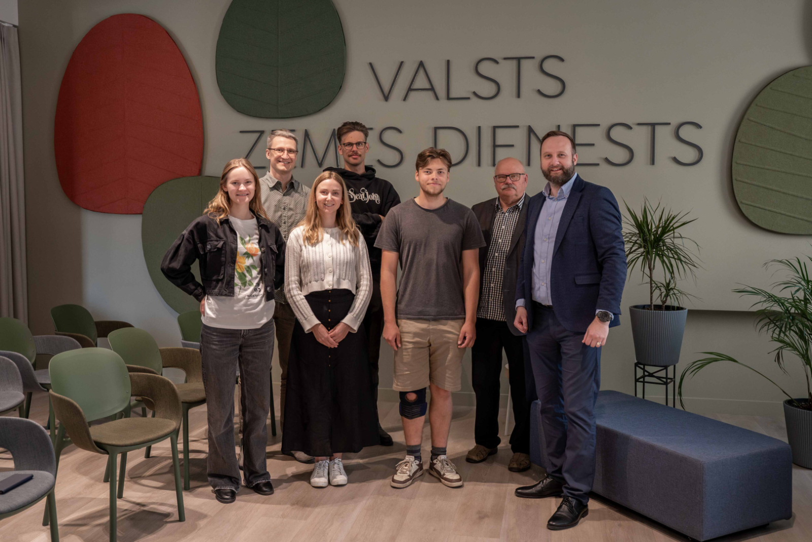 Tikšanās dalībnieku foto Valsts zemes dienesta Kadastra zālē, kur vairāki cilvēki stāv rindā pie sienas ar organizācijas nosaukumu.