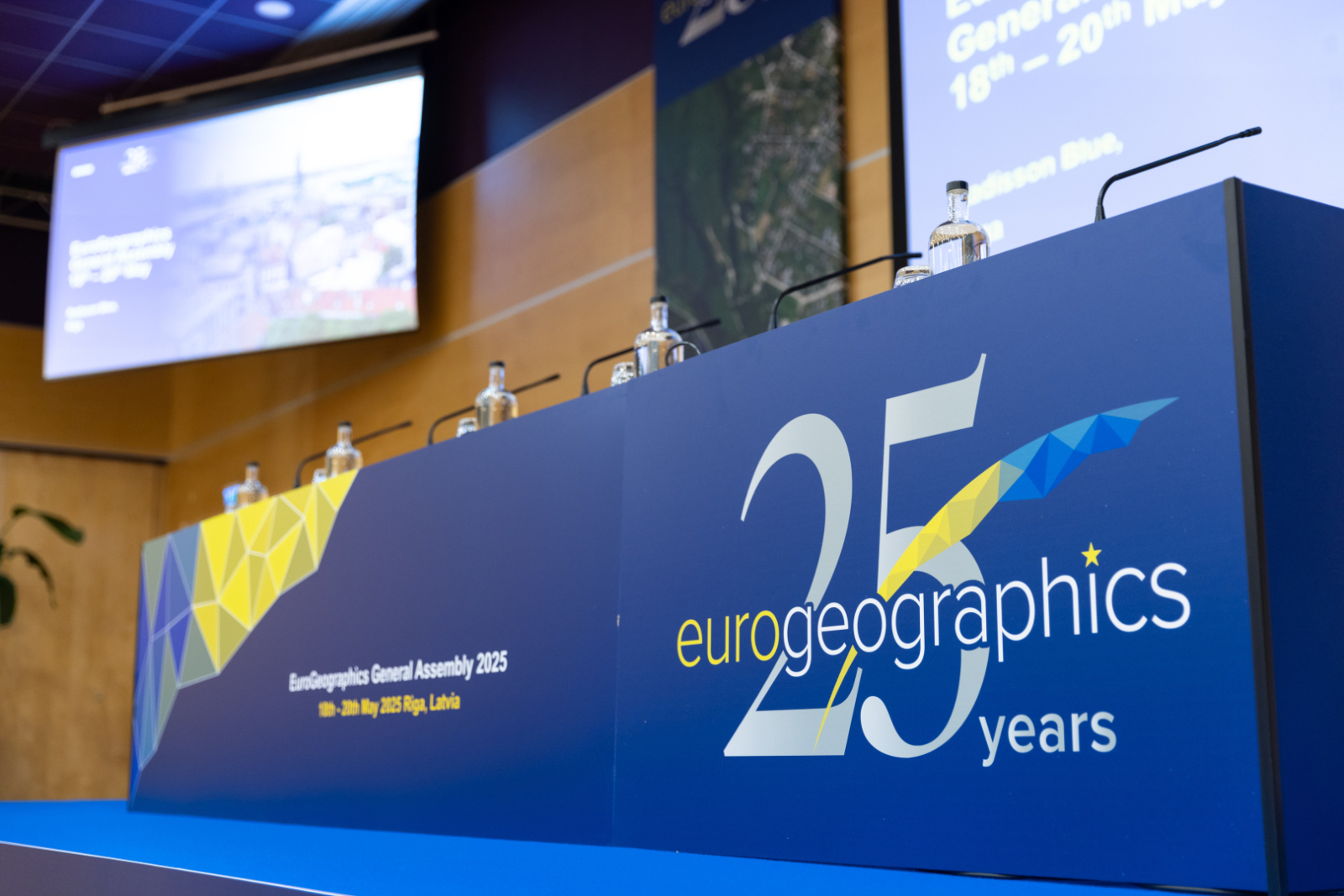 Konferences skatuve ar uzrakstu 'Eurogeographics 25 years' un ekrāniem, kas informē par EuroGeographics Ģenerālo asambleju Rīgā 2025. gadā.