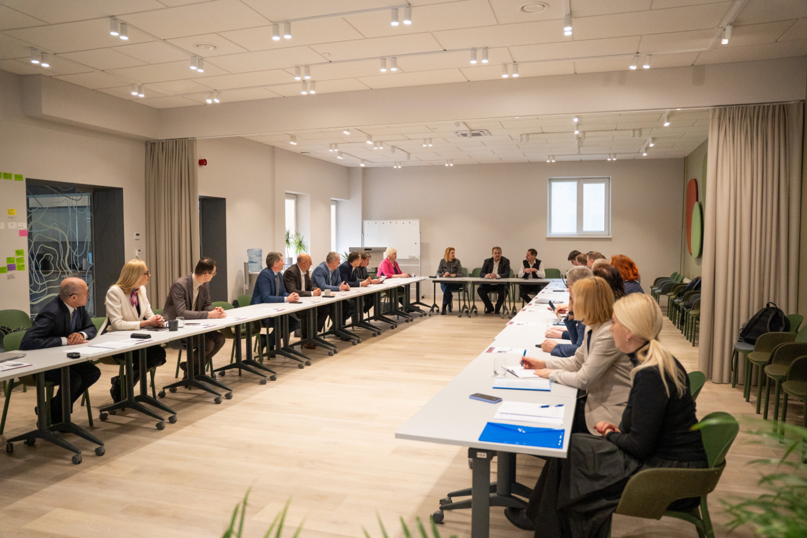 Gaiša sanāksmju telpa, kur pie divām galdų rindām profesionālā darba apspriedē piedalās pārstāvji no Valsts zemes dienesta un Saeimas Valsts pārvaldes un pašvaldības komisijas. Priekšgalā sēž prezentētāji, telpā moderns apgaismojums un neitrāls interjers.