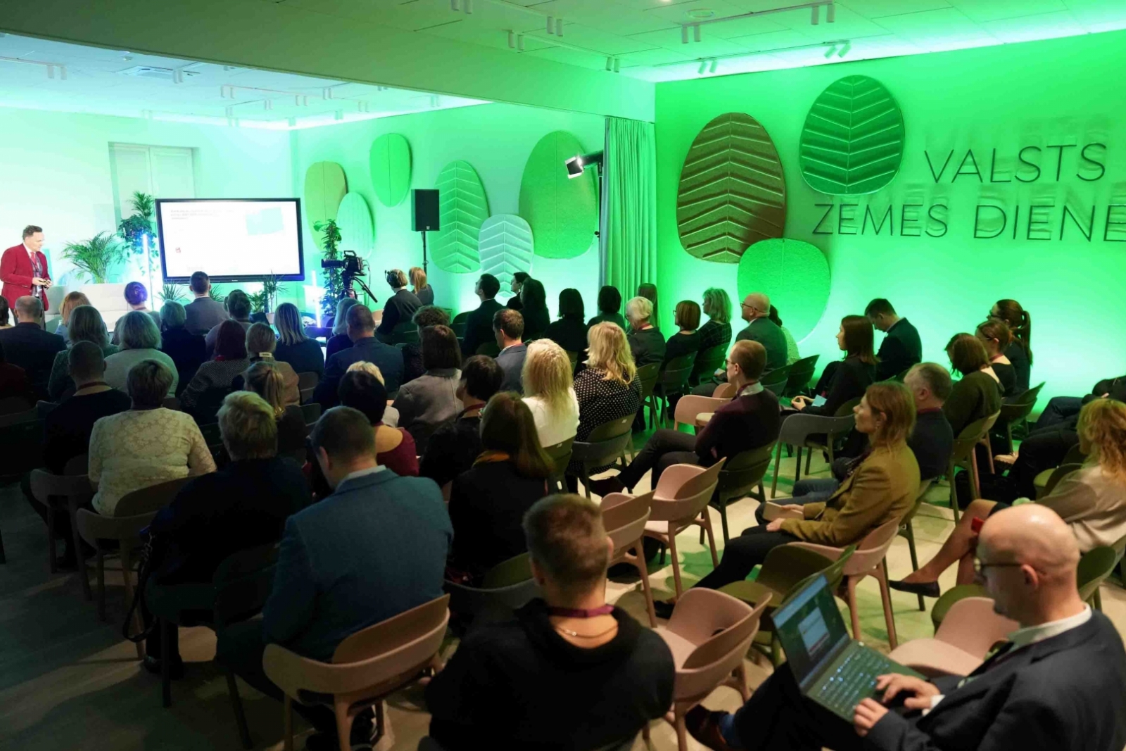 Valsts zemes dienesta Kadastra zāles foto, kurā notiek prezentācija par aktualitātēm. Dalībnieki sēž rindās, klausoties runātāju pie ekrāna. Telpu izgaismo spilgta zaļā gaisma un sienas rotā stilizēti koku lapu paneļi ar VZD nosaukumu, radot dinamisku un mūsdienīgu vidi.