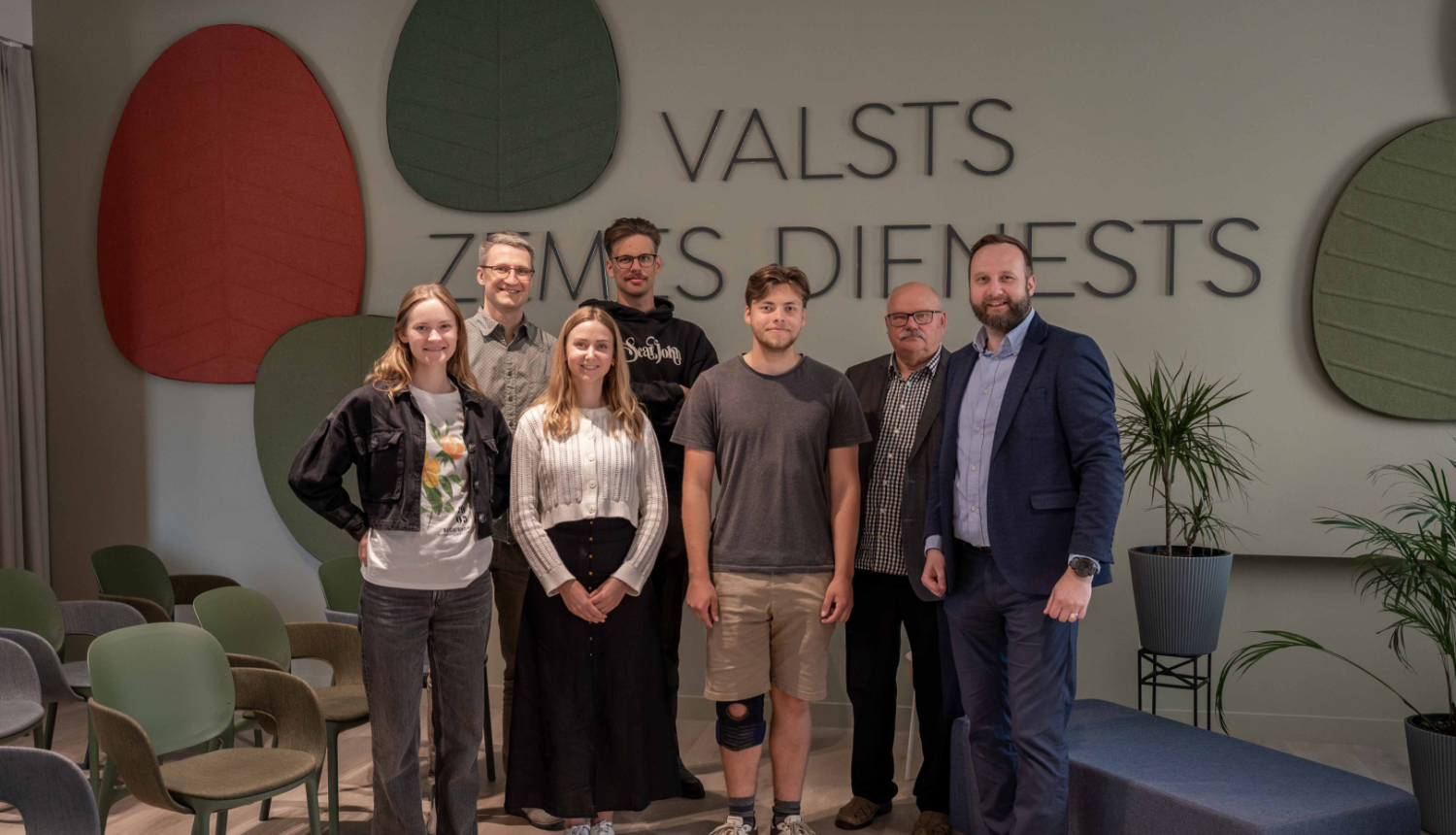 Tikšanās dalībnieku foto Valsts zemes dienesta Kadastra zālē, kur vairāki cilvēki stāv rindā pie sienas ar organizācijas nosaukumu.