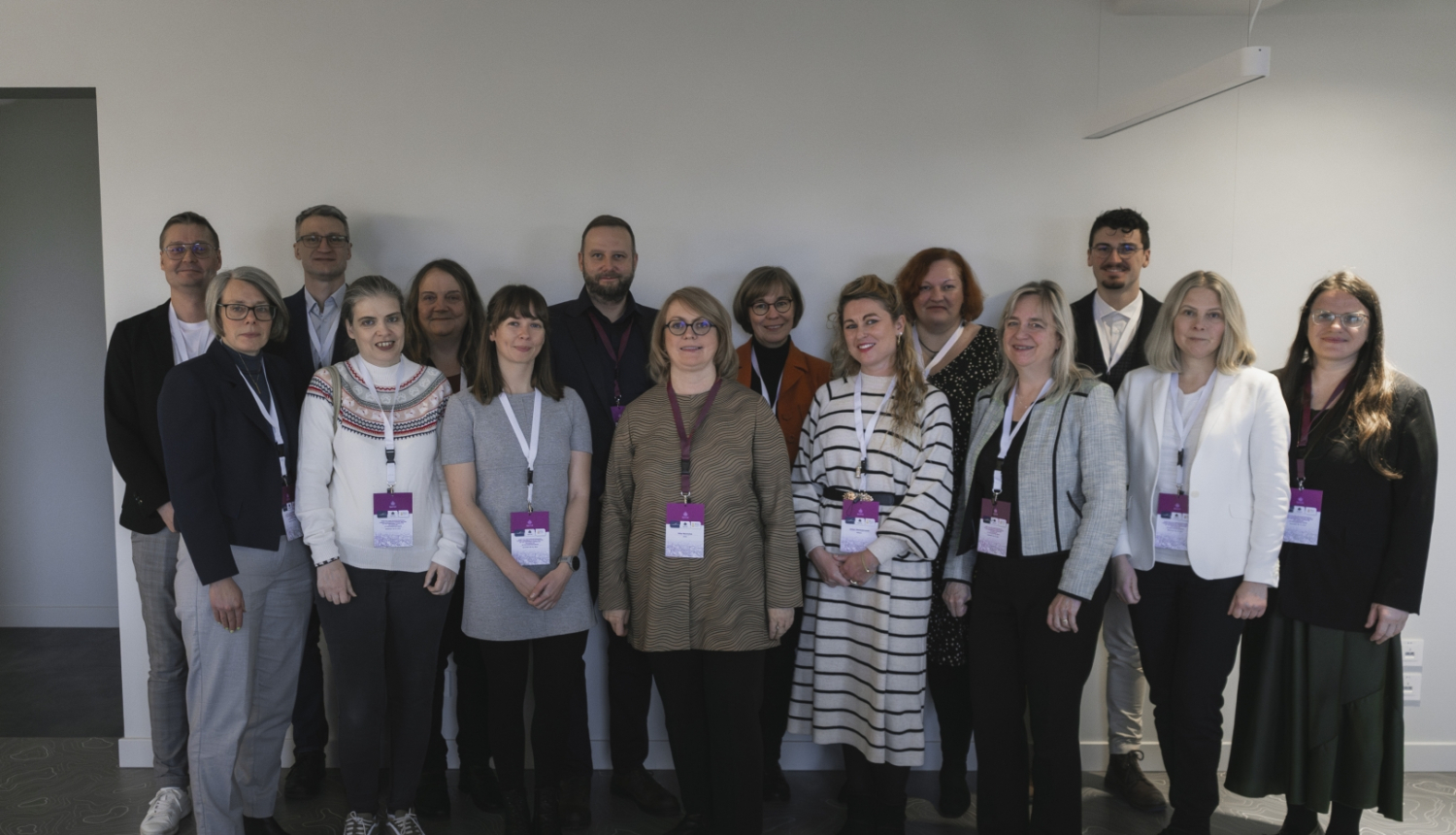 Konferences dalībnieku foto gaišā telpā, kurā redzami dažādu iestāžu pārstāvji. Visi dalībnieki stāv rindās pie sienas, valkā pasākuma identifikācijas kartes un oficiālu vai biznesa ikdienas apģērbu, radot organizētu un formālu noskaņu.