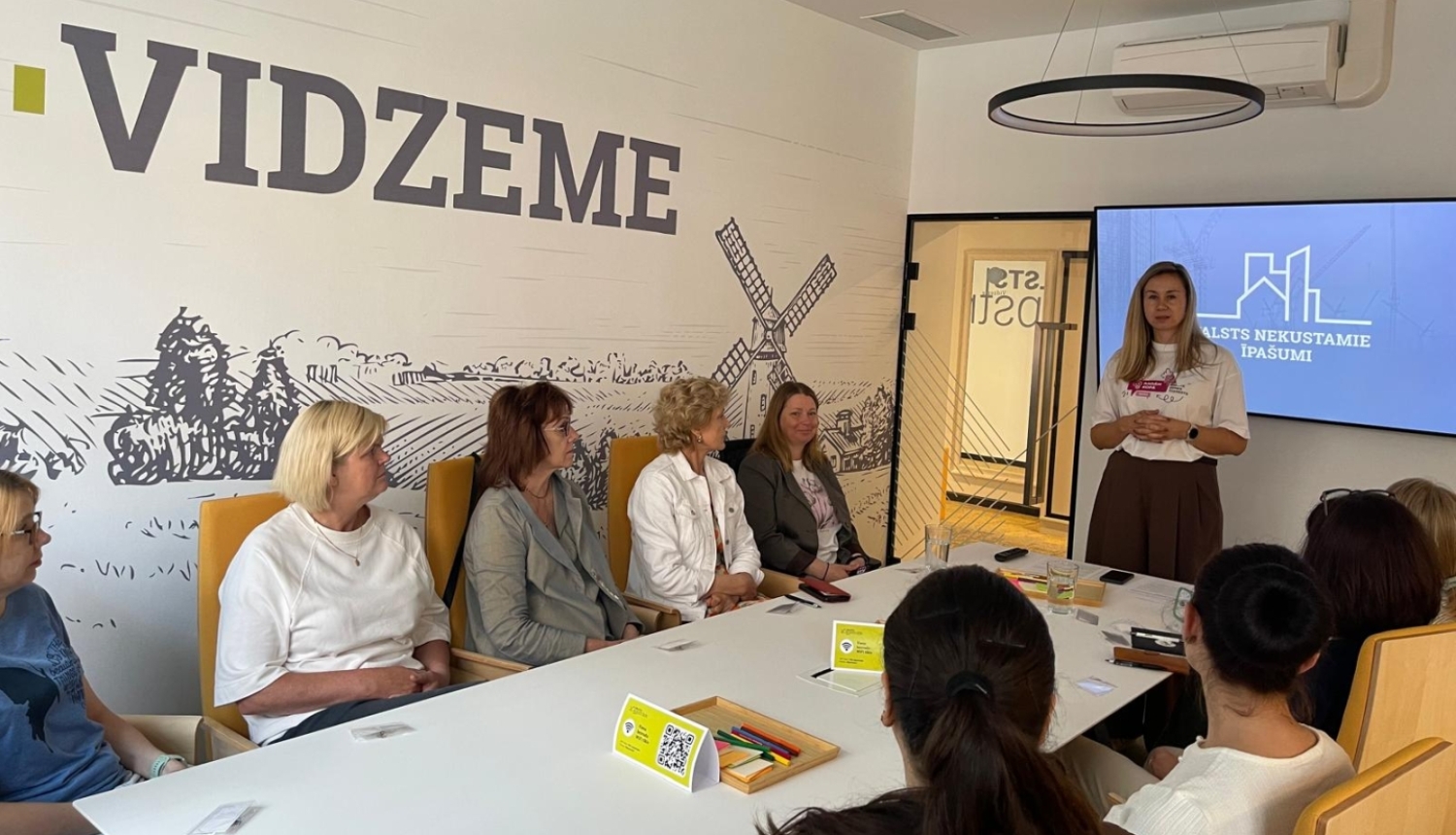 Telpā ar uzrakstu pie sienas "Vidzeme" pie galda sēž vairāki cilvēki
