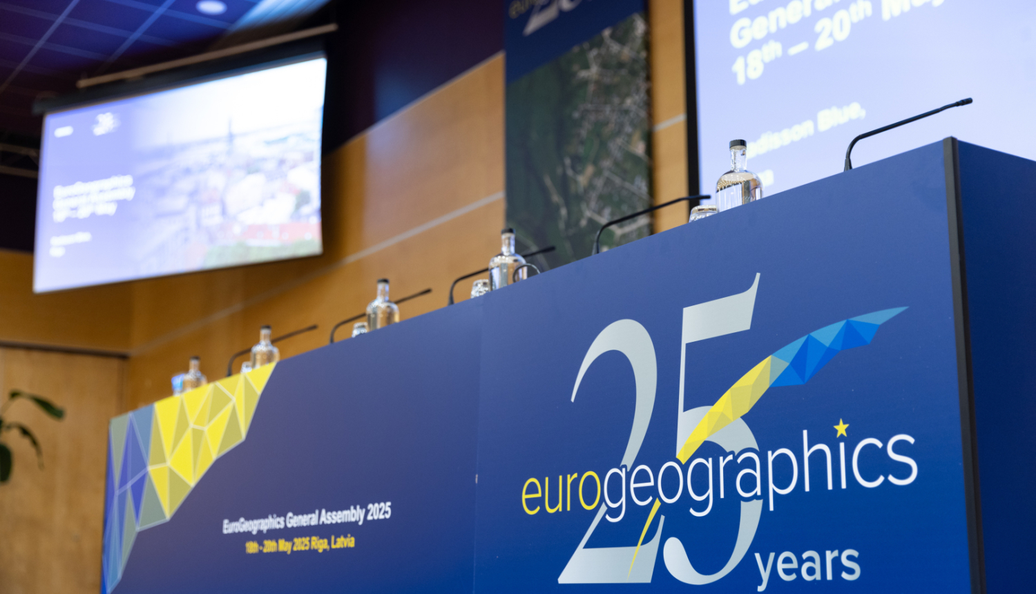 Konferences skatuve ar uzrakstu 'Eurogeographics 25 years' un ekrāniem, kas informē par EuroGeographics Ģenerālo asambleju Rīgā 2025. gadā.