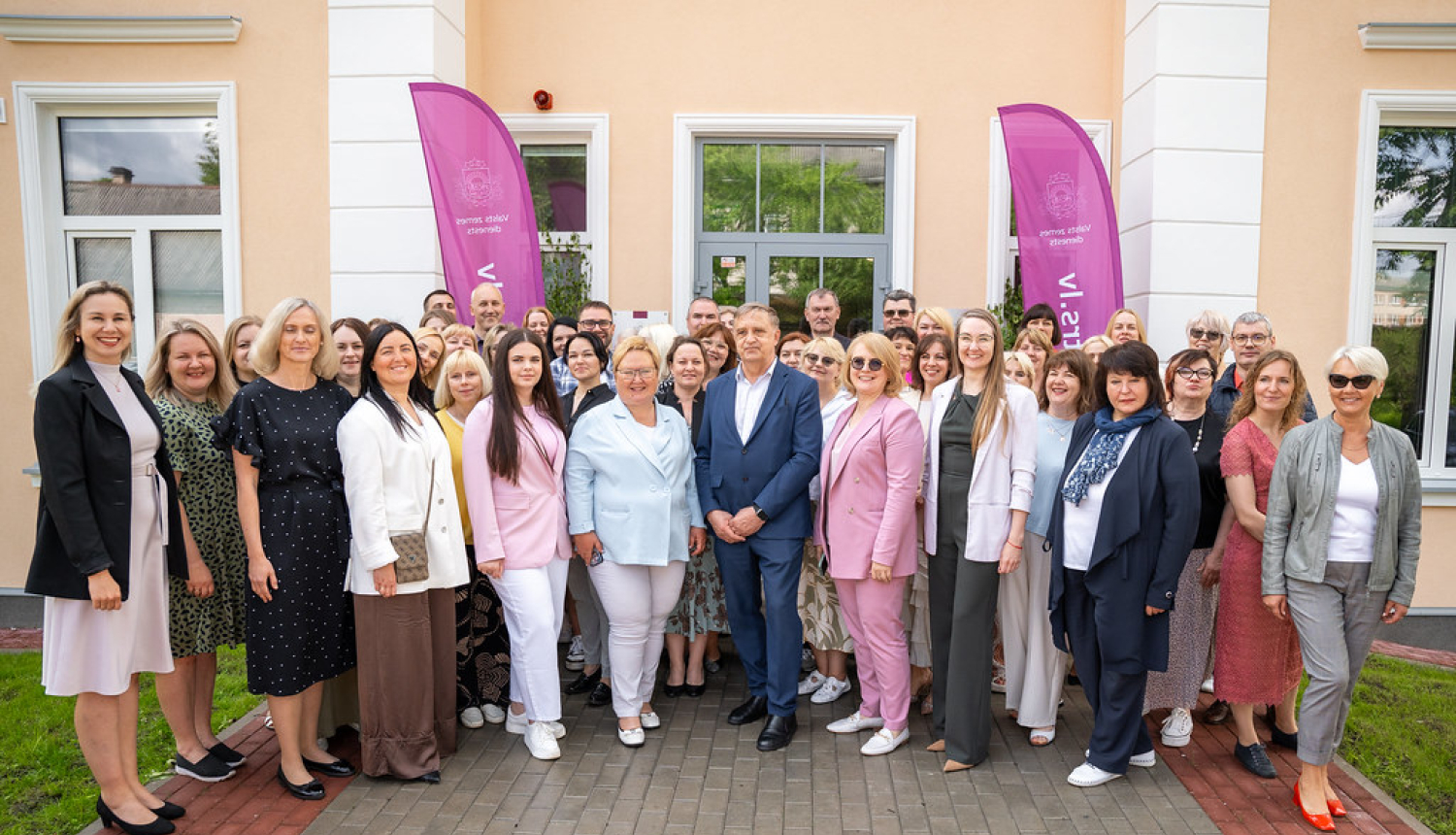 Pasākuma dalībnieku kopīga fotogrāfija pie Rēzeknes biroja ieejas, fonā divi violeti Valsts zemes dienesta karogi