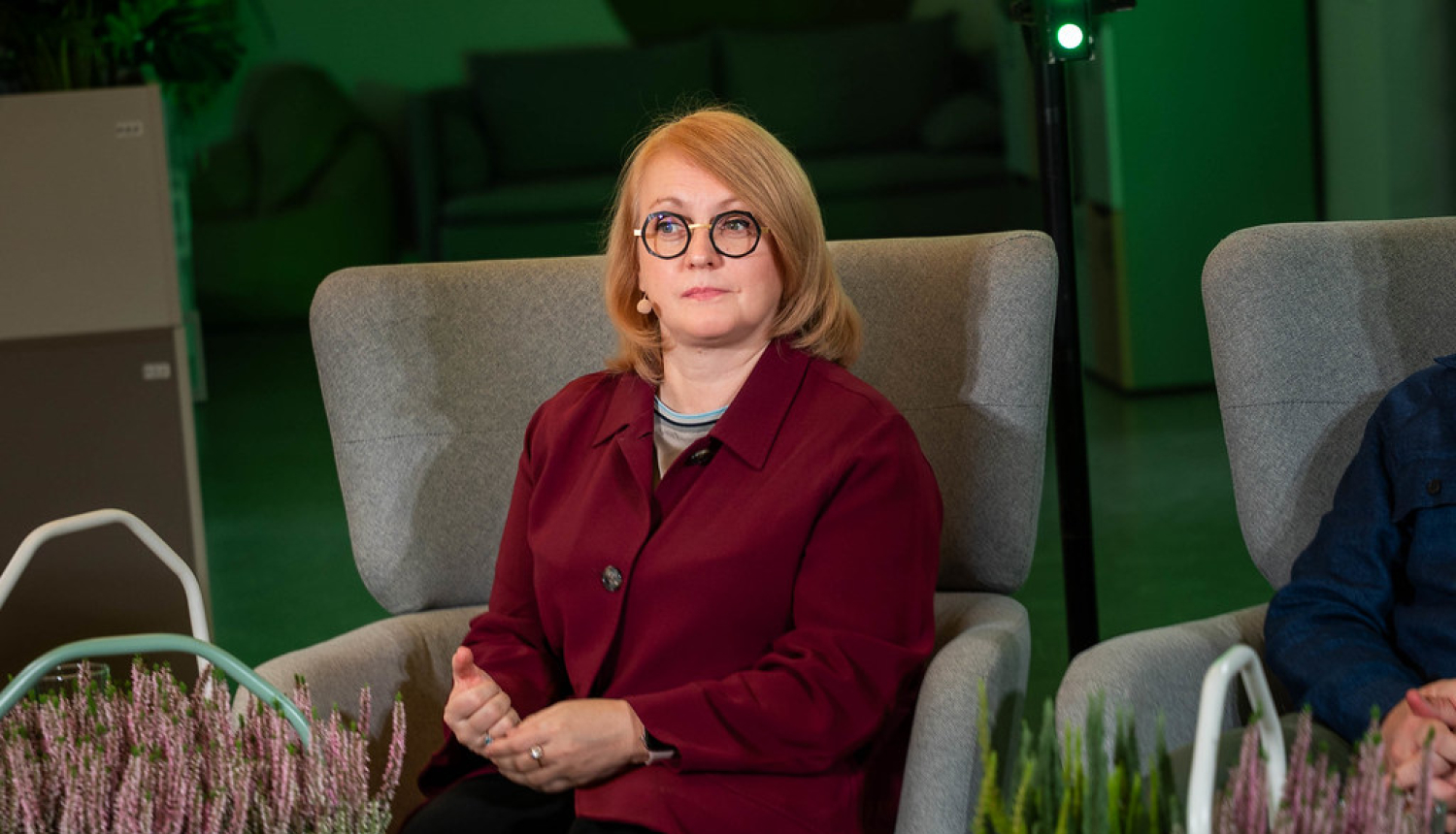 Valsts zemes dienesta ģenerāldirektore sēž krēslā , tērpusies tumši sarkanā žaketē. Priekšplānā redzami dekoratīvi augi, bet fonā — gaišs interjers ar dīvāniem un apgaismojumu.