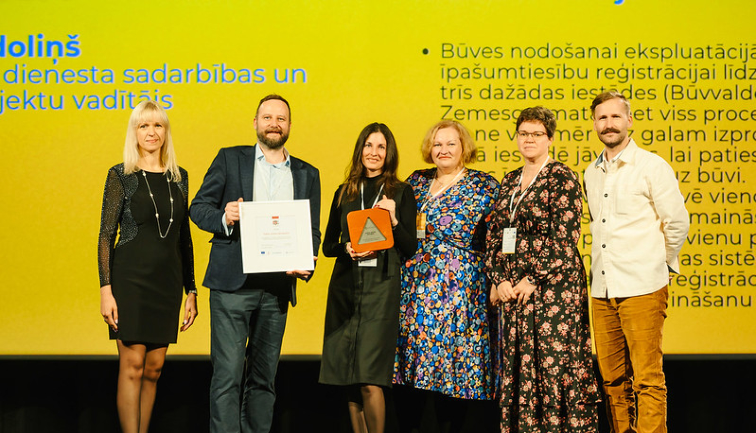 Sešu cilvēku grupa stāv skatuves priekšā pasākumā, rokās turot apbalvojumus un sertifikātu. Fonā redzams liels ekrāns ar dzeltenu prezentācijas slaidu un tekstu par sadarbību un būvju reģistrācijas procesu.