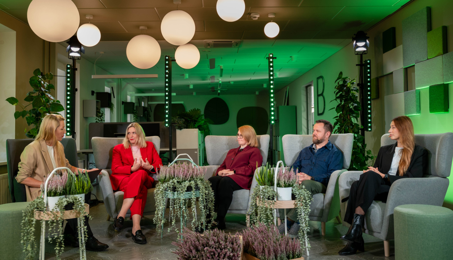 Paneļdiskusijas dalībnieku foto telpā ar zaļiem gaismas akcentiem. Dalībnieki sēž mīkstos krēslos puslokā, starp viņiem izvietoti dekoratīvi augi. Fonā redzams moderna interjera dizains ar griestu lampām, sienu paneļiem un telpaugiem.