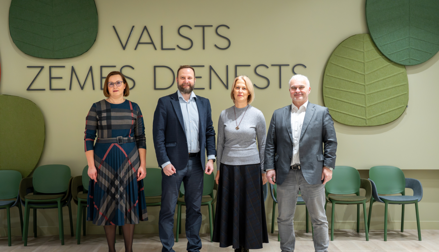 Tikšanās dalībnieki stāv rindā pie sienas ar uzrakstu “Valsts zemes dienests”. Fons veidots gaišos toņos ar lielām dekoratīvām lapu formas plāksnēm un rindā novietotiem krēsliem.