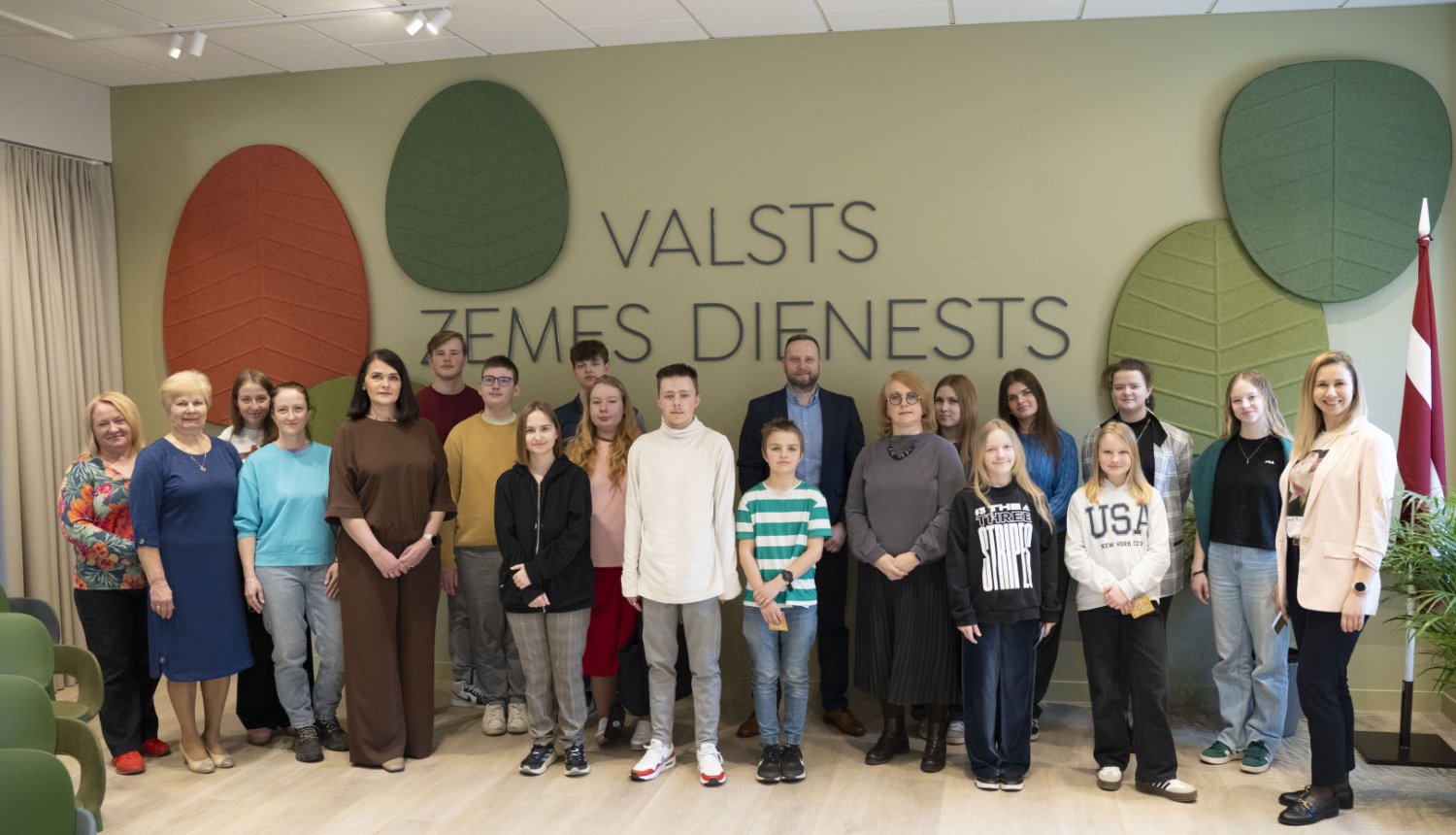 Grupas foto Valsts zemes dienesta telpā pie sienas ar VZD nosaukumu un stilizētiem koku lapu dekoriem. Priekšplānā redzami Valsts zemes dienesta darbinieki un jaunieši, kuri piedalās "Ēnu diena" pasākumā. Fonā redzams arī Latvijas karogs un moderna, gaiša telpa.