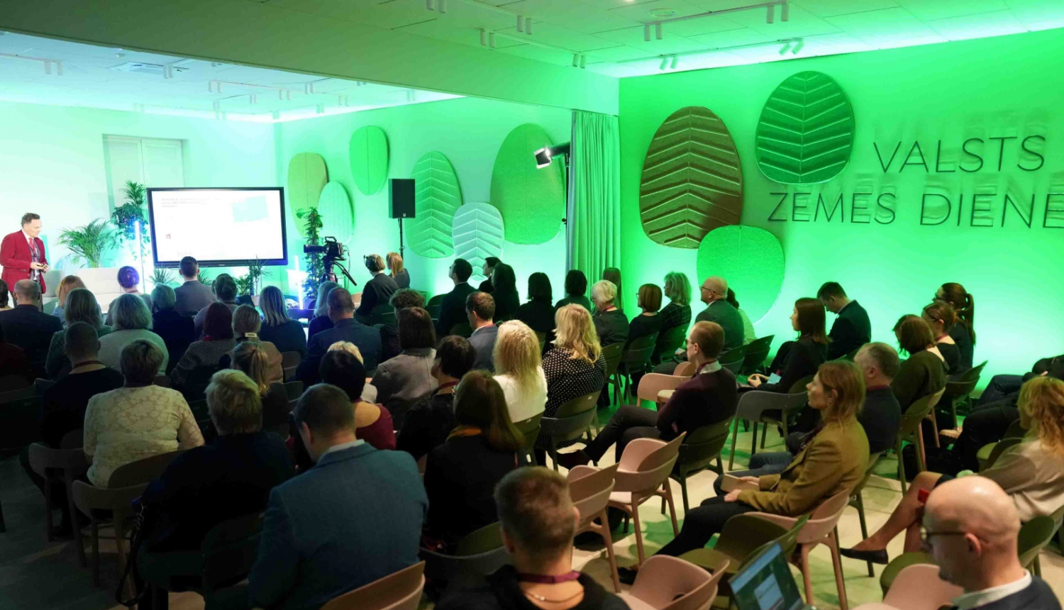 Valsts zemes dienesta Kadastra zāles foto, kurā notiek prezentācija par aktualitātēm. Dalībnieki sēž rindās, klausoties runātāju pie ekrāna. Telpu izgaismo spilgta zaļā gaisma un sienas rotā stilizēti koku lapu paneļi ar VZD nosaukumu, radot dinamisku un mūsdienīgu vidi.