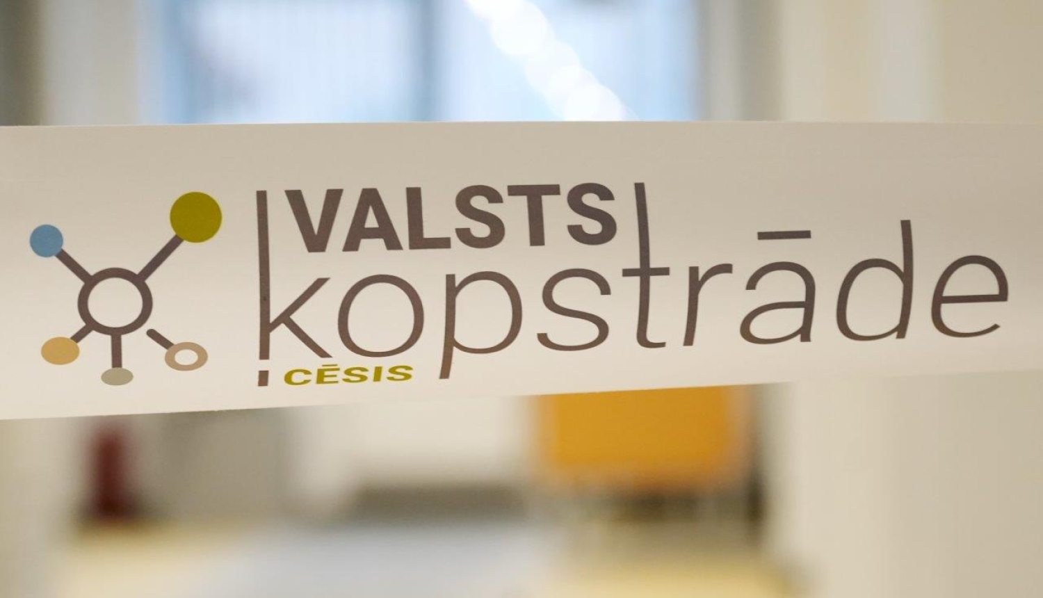 Tuvplāna attēls ar balti laminētu lentu, uz kuras redzams “Valsts kopstrāde Cēsis” vizuālais apzīmējums un stilizēta sadarbības tīkla ikona. Fons ir gaišs biroja gaiteņa interjers ar izpludinātām formām, kas izceļ priekšplānā redzamo zīmolu.