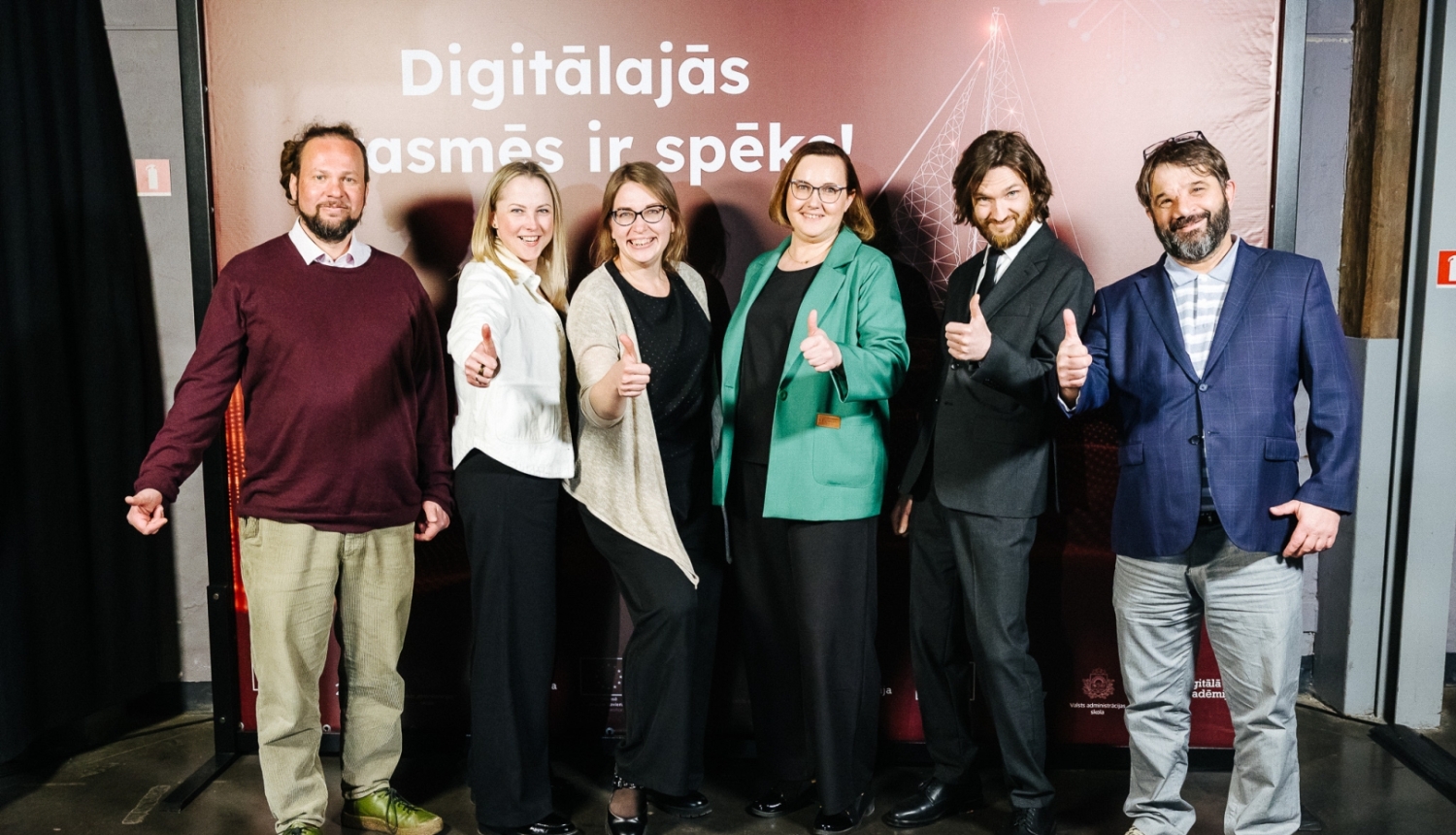 Sešu cilvēku grupa smaida un rāda paceltus īkšķus uz pasākuma stenda fona ar uzrakstu "Digitālajās prasmēs ir spēks!"