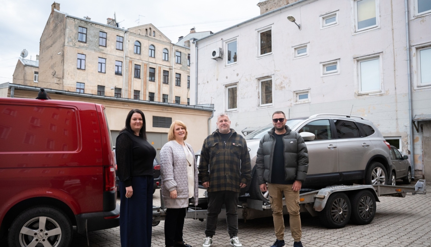Četri cilvēki stāv pagalmā pie sarkanas furgona un piekabes ar sudraba apvidus automašīnu; fonā redzamas vecākas daudzdzīvokļu ēkas.