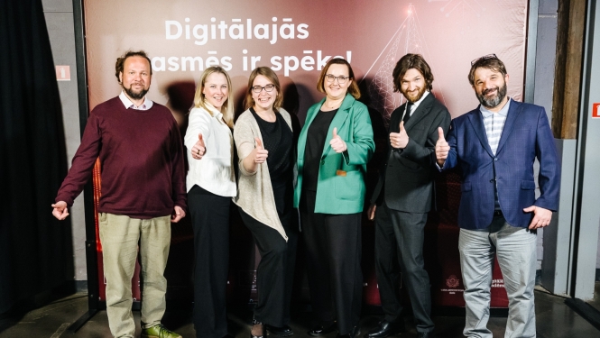 Sešu cilvēku grupa smaida un rāda paceltus īkšķus uz pasākuma stenda fona ar uzrakstu "Digitālajās prasmēs ir spēks!"