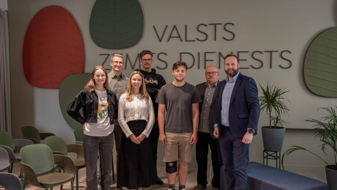 Tikšanās dalībnieku foto Valsts zemes dienesta Kadastra zālē, kur vairāki cilvēki stāv rindā pie sienas ar organizācijas nosaukumu.