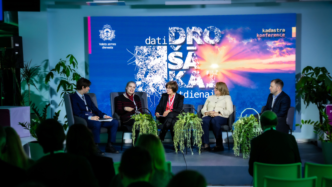 Paneļdiskusijas skats uz skatuves, kur pieci dalībnieki sēž krēslos un runā par datu nozīmi drošības un krīžu vadībā. Fonā redzams liels ekrāns ar konferences vizuālo noformējumu “Dati drošākai rītdienai”, bet priekšplānā – auditorijas silueti.