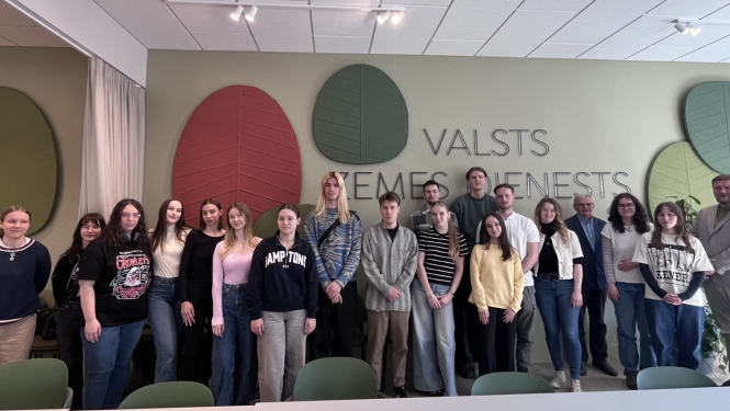 Grupas foto Valsts zemes dienesta telpās: vairāki jaunieši un pieaugušie stāv rindā pretī kamerai. Fonā redzams uzraksts “Valsts zemes dienests” un dekoratīvi lapu formas sienas elementi. Telpa ir gaiša, ar krēsliem priekšplānā.