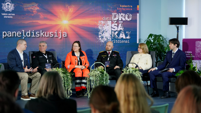 Uz skatuves norisinās paneļdiskusija par drošības un krīžu vadību. Seši dalībnieki sēž krēslos, tostarp operatīvo dienestu pārstāvji uniformās un spilgti oranžā darba tērpā. Fonā redzams liels ekrāns ar uzrakstiem “Paneļdiskusija” un “Dati drošākai rītdienai”, bet priekšplānā – auditorijas silueti.