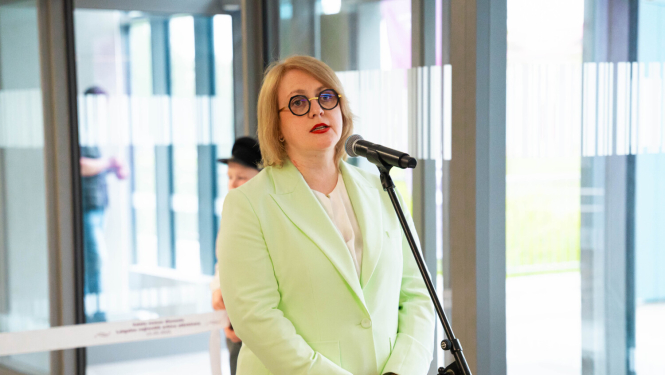 Attēlā Valsts zemes dienesta ģenerāldirektore Vita Narnicka, tērpusies gaiši zaļā žaketē, uzrunā klātesošos pie mikrofona modernā ēkas foajē ar stiklotām sienām