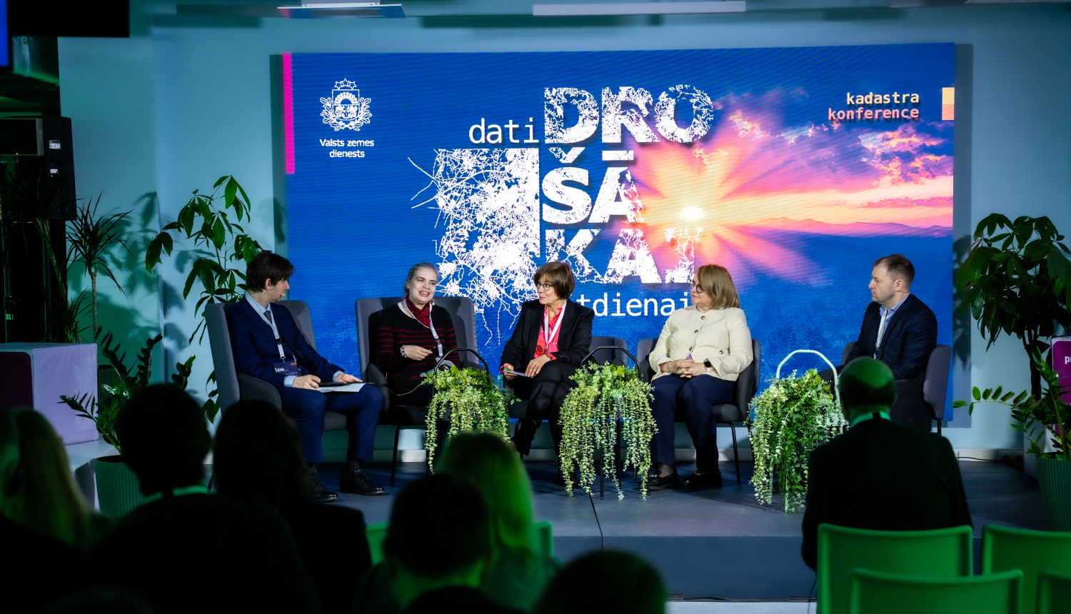 Paneļdiskusijas skats uz skatuves, kur pieci dalībnieki sēž krēslos un runā par datu nozīmi drošības un krīžu vadībā. Fonā redzams liels ekrāns ar konferences vizuālo noformējumu “Dati drošākai rītdienai”, bet priekšplānā – auditorijas silueti.