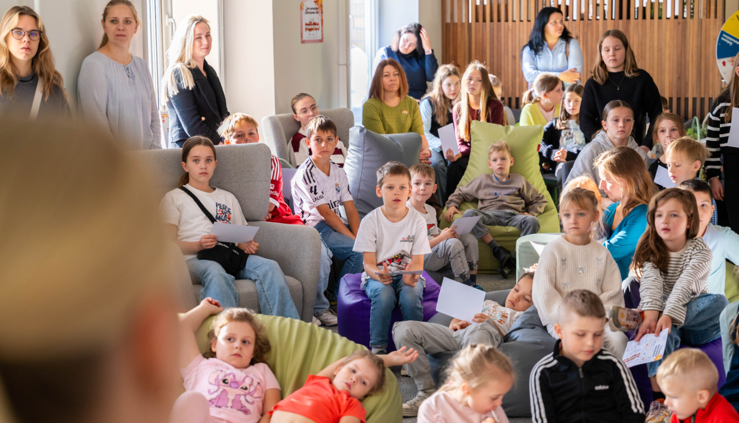 Pasākuma dalībnieki pulcējušies gaišā telpā uz kopīgu nodarbību. Daži sēž uz pufiem vai dīvāniem, citi stāv, rokās turot papīrus. Telpā redzams silts apgaismojums un koka interjera elementi.