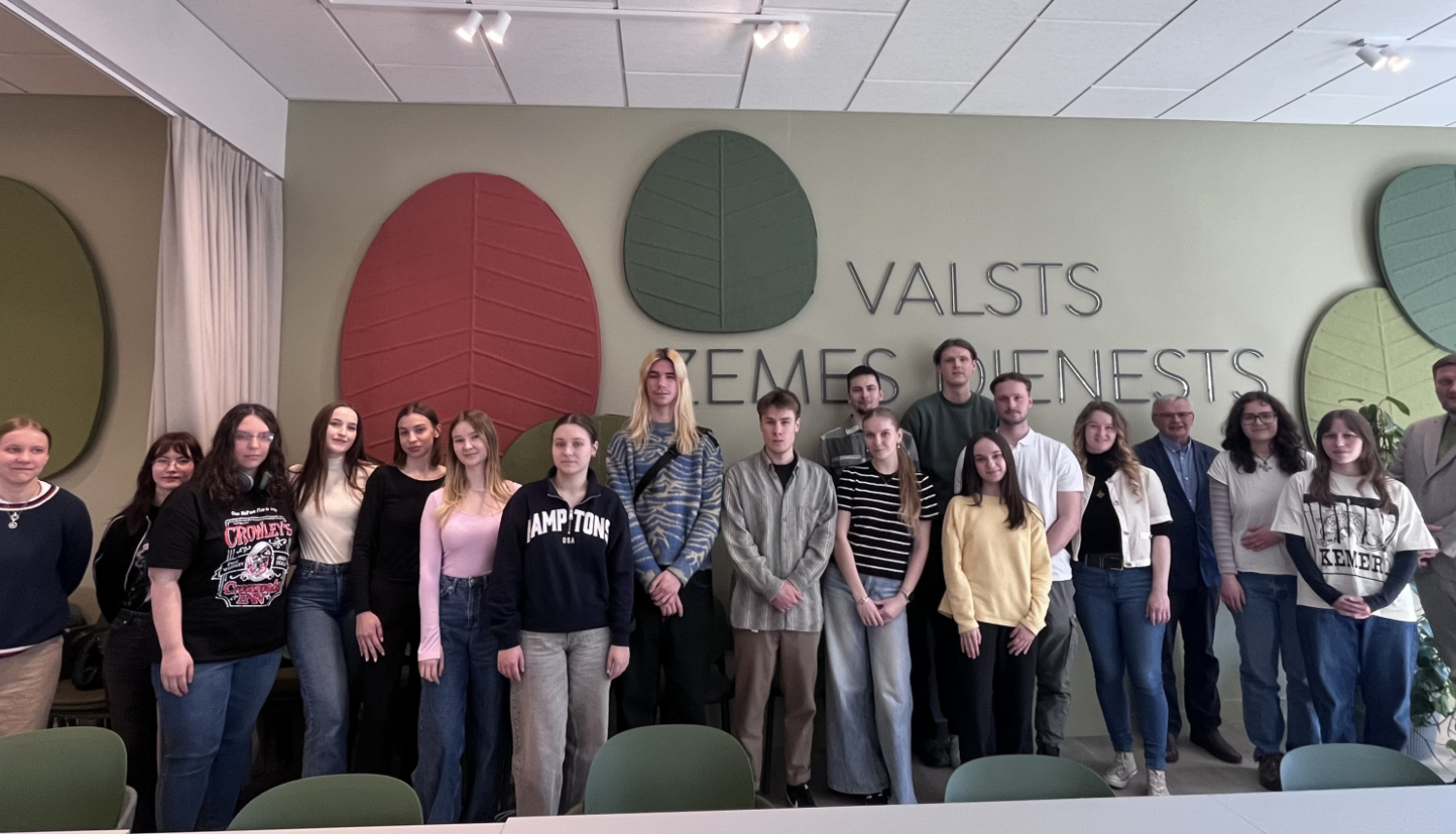 Grupas foto Valsts zemes dienesta telpās: vairāki jaunieši un pieaugušie stāv rindā pretī kamerai. Fonā redzams uzraksts “Valsts zemes dienests” un dekoratīvi lapu formas sienas elementi. Telpa ir gaiša, ar krēsliem priekšplānā.