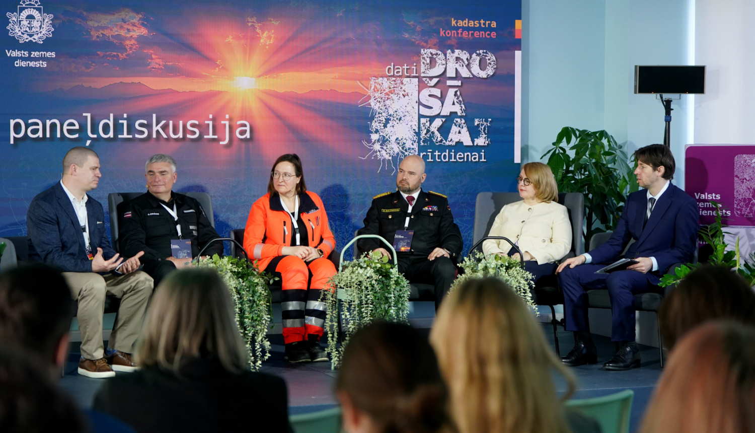 Uz skatuves norisinās paneļdiskusija par drošības un krīžu vadību. Seši dalībnieki sēž krēslos, tostarp operatīvo dienestu pārstāvji uniformās un spilgti oranžā darba tērpā. Fonā redzams liels ekrāns ar uzrakstiem “Paneļdiskusija” un “Dati drošākai rītdienai”, bet priekšplānā – auditorijas silueti.