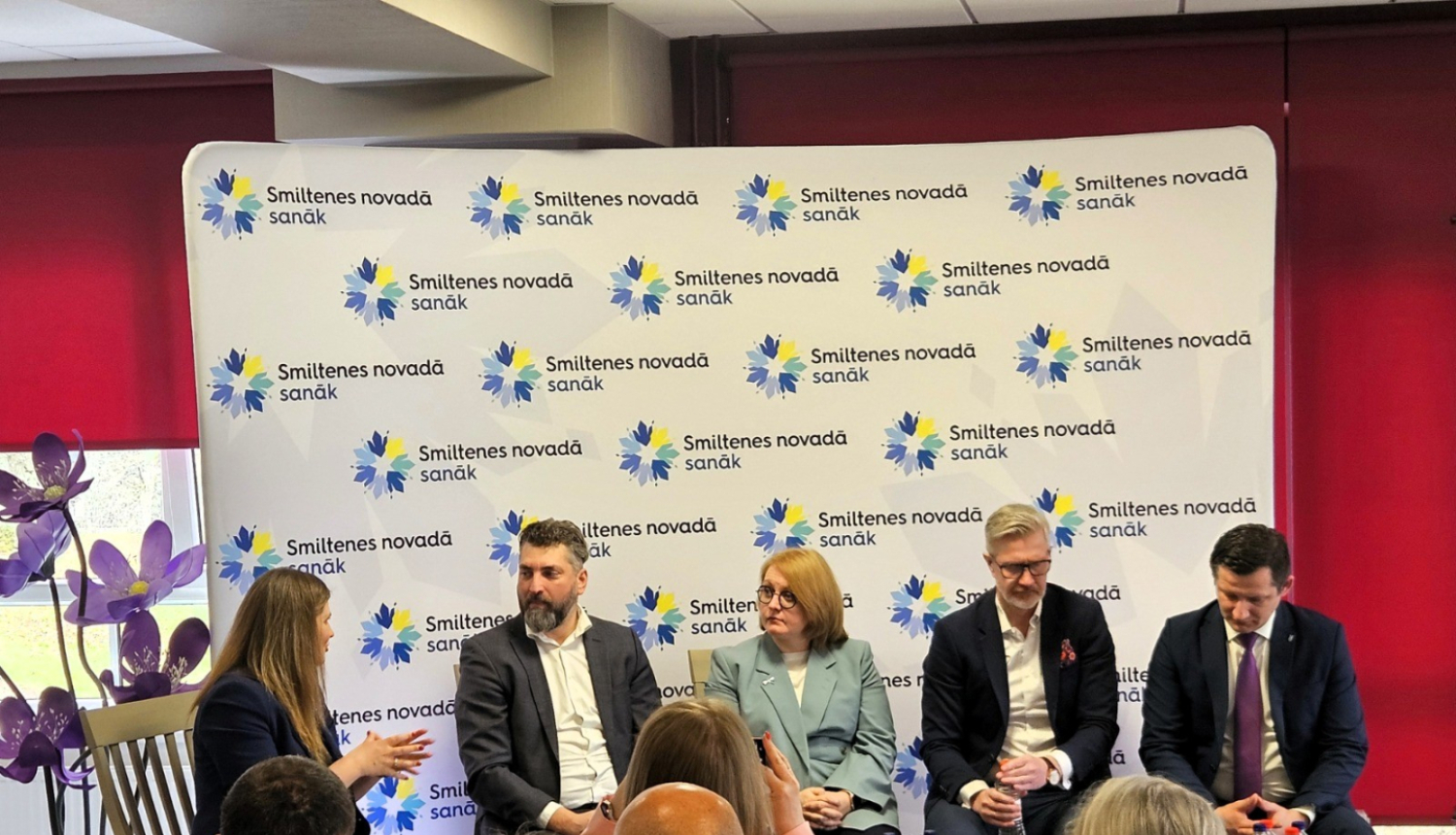 Attēlā redzama LTRK paneļdiskusija. Piecu personu grupa sēž auditorijas priekšā, fonā stends ar uzrakstu ‘Smiltenes novadā satiek’. Klausītāji sēž pirmajā plānā, bet runātāji diskutē.