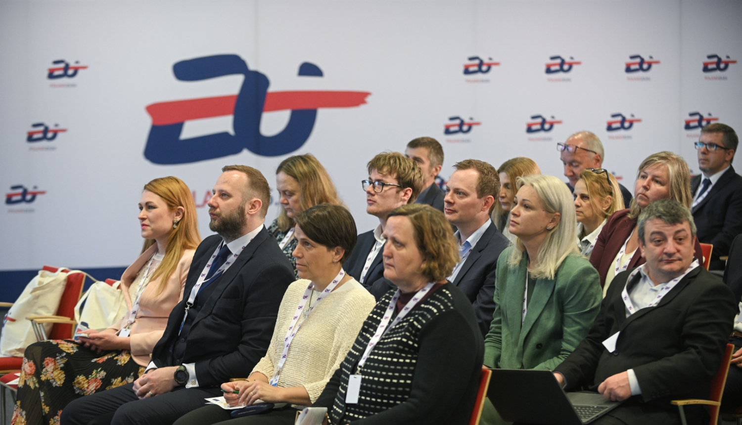 Konferences dalībnieki sēž auditorijā un klausās prezentāciju, fonā redzams pasākuma grafiskais noformējums ar Eiropas kadastra tematiku.